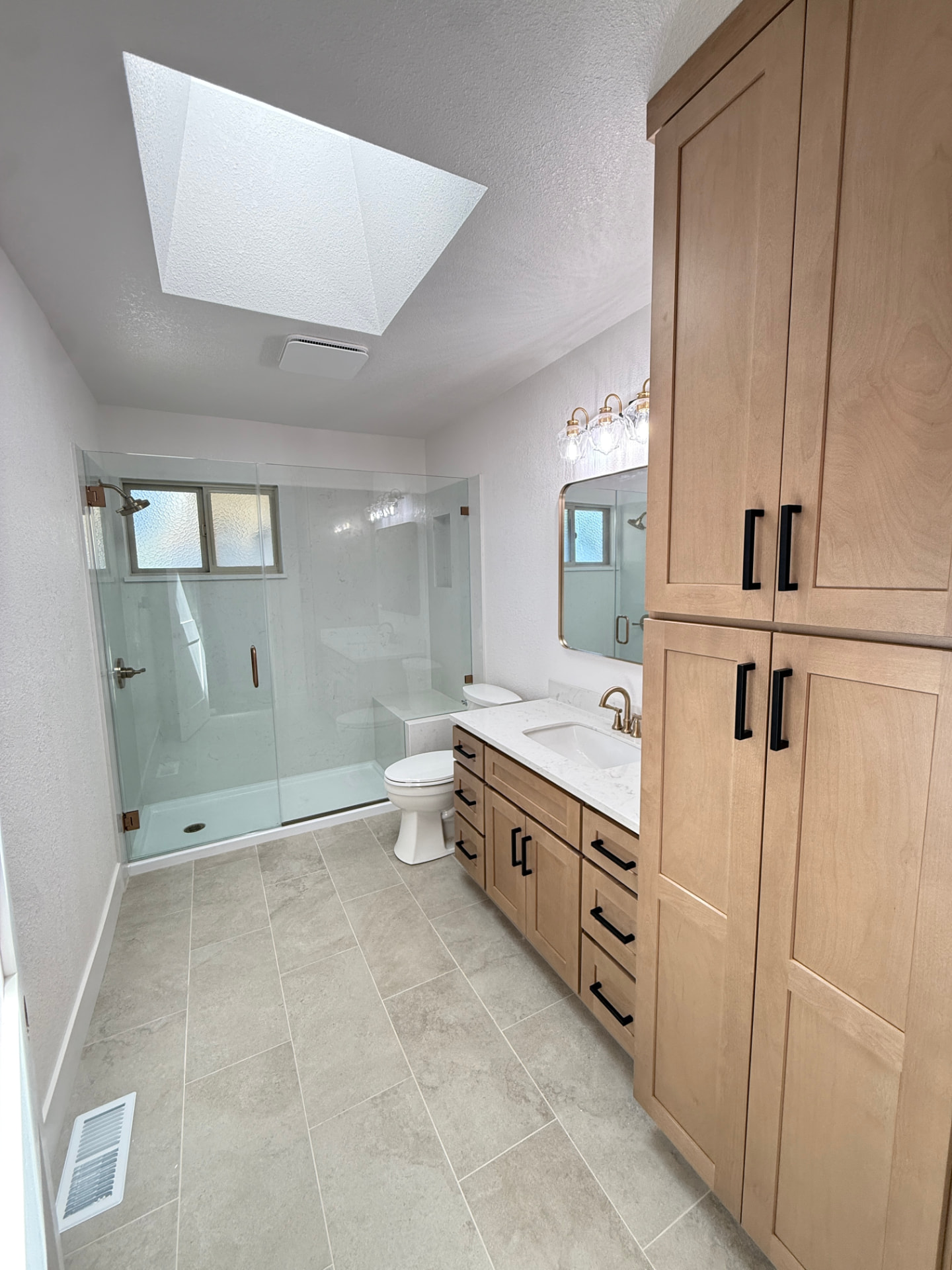House Doctor bathroom remodel with a wood vanity, brass faucet, full-height linen tower, and a skylit glass shower
